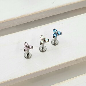 May include: Three surgical steel body jewelry pieces with a three-pronged design. Each piece has a different color of crystal: pink, clear, and blue.