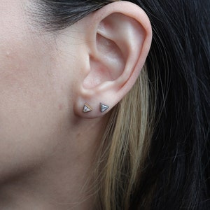May include: A close-up of a person's ear with two triangle-shaped earrings. The earring on the left is gold with a clear stone, and the earring on the right is silver with a clear stone.