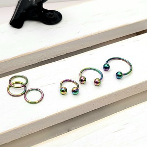 May include: A set of five rainbow-colored metal body jewelry pieces. Three are horseshoe-shaped with ball ends, and two are circular rings.