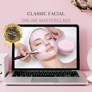 May include: A laptop screen displays a Classic Facial Online Masterclass with a person receiving a pink facial mask application. A gold accredited seal is attached to the left side of the screen. The laptop has a silver keyboard and a black frame.