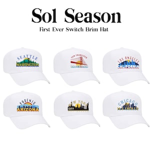 以下が含まれることがあります： 前面に異なる都市の街並みと名前が刺繍された、白いベースボールキャップが6つ。キャップはすべて白で、つばとストラップは黒。掲載されている都市は、シアトル、サンフランシスコ、ロサンゼルス、フェニックス、ニューヨークシティ、シカゴです。