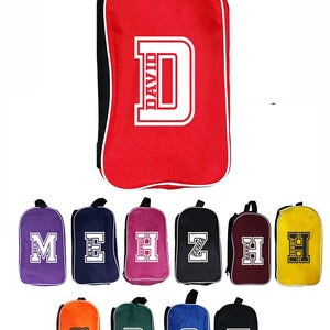 May include: A collection of personalised drawstring bags in various colours, including red, purple, navy, pink, black, orange, green, blue, and yellow. Each bag has a large initial and name printed in white.