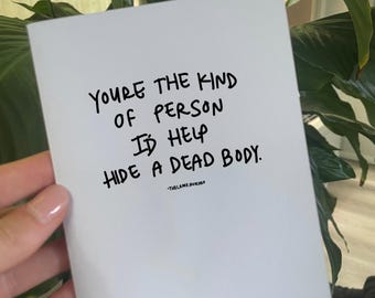 May include: A white card with black handwritten text that reads, "YOU'RE THE KIND OF PERSON I'D HELP HIDE A DEAD BODY." The card is held in front of a green plant with white flowers.