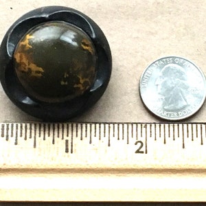 May include: A black button with a green and gold marbled design. The button is about the size of a US quarter.