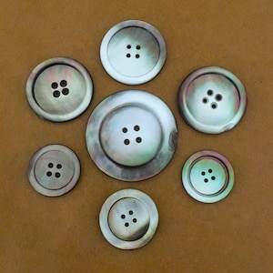 May include: Seven iridescent mother-of-pearl buttons of varying sizes are arranged on a brown surface. Each button has four holes for sewing. The buttons have a pearlescent sheen with hints of pink, green, and blue.