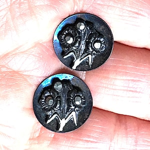 May include: Two antique black buttons with a floral bouquet design. Each button features a raised floral pattern with three flower heads and a stem. The buttons are round and have a subtle iridescent sheen.