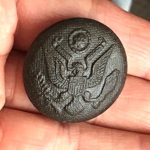 May include: A dark brown, round button with a textured surface. The button features a detailed embossed design of an eagle with a shield and stars, a classic emblem. This vintage button is likely from a uniform or military garment.
