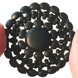 May include: A black, circular brooch with a central, smooth, round element. The brooch is surrounded by a pattern of small, oval and round beads, creating a textured, openwork design. The brooch is held between two fingers.
