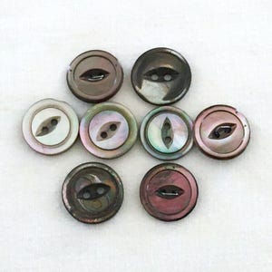 May include: A collection of eight round buttons with a mother-of-pearl appearance. The buttons vary in color, including shades of brown, gray, and pink. Each button has two small holes for sewing and a decorative, eye-shaped design.