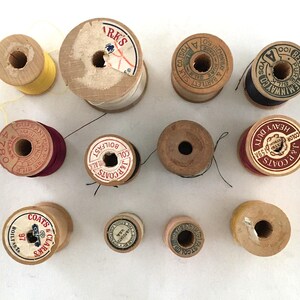 May include: A collection of vintage wooden spools of thread in various colors, including yellow, white, black, and red. Some spools have labels with text like "J&P Coats" and "ARK'S". The spools are arranged on a white surface.