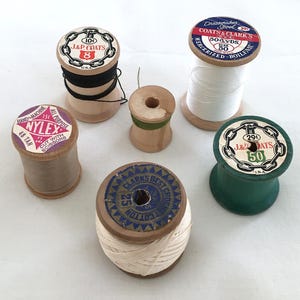 May include: A collection of vintage wooden spools of thread in various colors, including black, white, tan, and green. The spools have labels with text such as "J&P Coats" and "Nylex". Some spools have thread still wound around them.