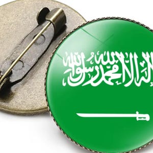 May include: A round pinback button featuring the flag of Saudi Arabia. The button has a green background with white Arabic script and a sword. The pin has a metal clasp on the back.