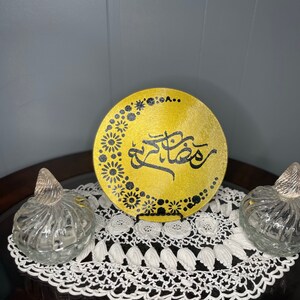 May include: A gold-colored decorative plate with black Arabic calligraphy and floral patterns. The plate is displayed on a white lace doily, flanked by two clear glass jars with decorative stoppers. The items are arranged on a dark wooden surface.