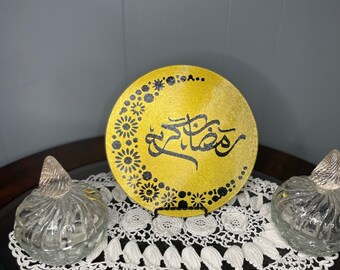 May include: A gold-colored decorative plate with black Arabic calligraphy and floral patterns. The plate is displayed on a white lace doily, flanked by two clear glass jars with decorative stoppers. The items are arranged on a dark wooden surface.