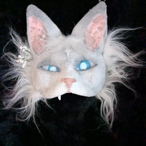 May include: A handcrafted cat mask in white and grey. The mask has large, pointed ears with pink inner details, striking blue eyes, and a pink nose. White faux fur frames the face, and small floral accents are placed on the ears.