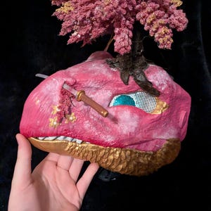 May include: A pink and gold mask with a textured surface, a small sword, and a mesh eye. The mask is adorned with a cluster of pink and gold flowers. The mask is held by a hand.