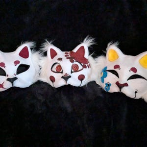 May include: Three white cat masks with different designs. One has a red slash, another has red accents, and the third has a blue flower and yellow ears. Each mask has black eye cutouts and white fur trim.