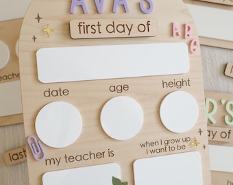 May include: Wooden first day of school sign with the name "AVA'S" in purple letters. The sign includes spaces for date, age, and height, with a red apple accent. Additional signs are visible.