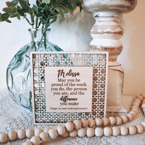 May include: Wooden plaque with the name "Melissa" and a message for a 20-year work anniversary. The plaque has a decorative border and is displayed with a vase and wooden beads.