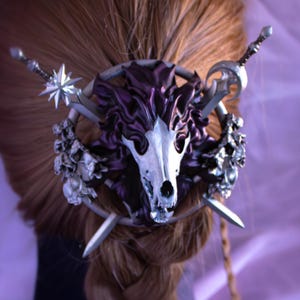 May include: A silver hair accessory featuring a skull design with purple accents, adorned with small flowers and crossed swords. The hair piece is set against a backdrop of braided brown hair and a purple background.
