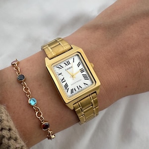 May include: A gold-toned rectangular wristwatch with a white face and black Roman numerals is worn on a wrist. A gold bracelet with colorful gemstones is also visible. The watch band and watch case are gold-colored.