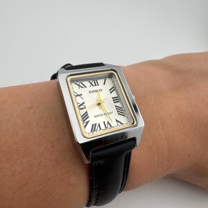 May include: A rectangular Casio watch with a silver-tone case, gold-tone accents, and a cream-colored face. The watch features Roman numeral hour markers and black leather band. The text "CASIO" and "WATER RESIST" are visible on the watch face.