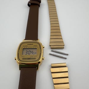 K&ouml;nnte beinhalten: Eine goldfarbene Casio Digitaluhr mit braunem Lederarmband, die die Uhrzeit 3:04:54 anzeigt. Das Bild zeigt auch ein abgenommenes goldenes Metallarmband und zwei Stifte, was auf eine Reparatur oder einen Armbandwechsel hindeutet.