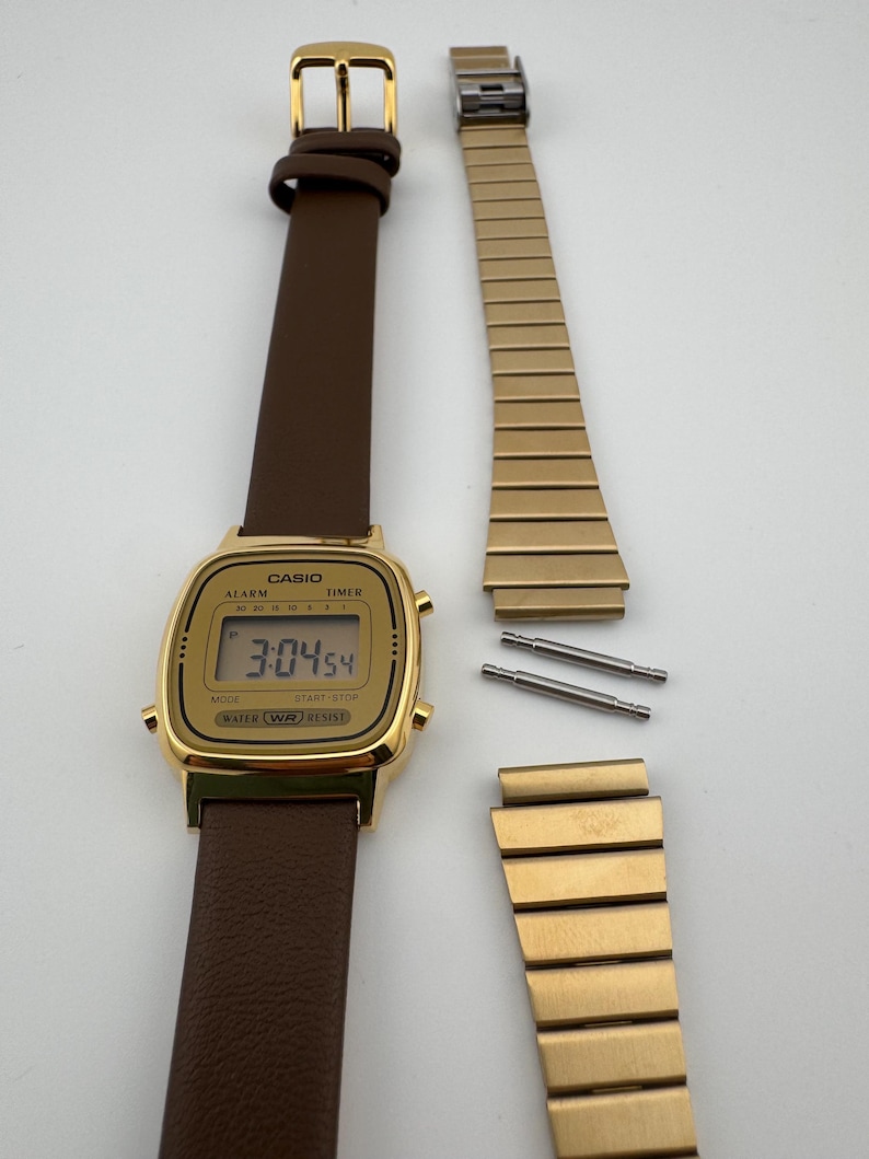 K&ouml;nnte beinhalten: Eine goldfarbene Casio Digitaluhr mit braunem Lederarmband, die die Uhrzeit 3:04:54 anzeigt. Das Bild zeigt auch ein abgenommenes goldenes Metallarmband und zwei Stifte, was auf eine Reparatur oder einen Armbandwechsel hindeutet.