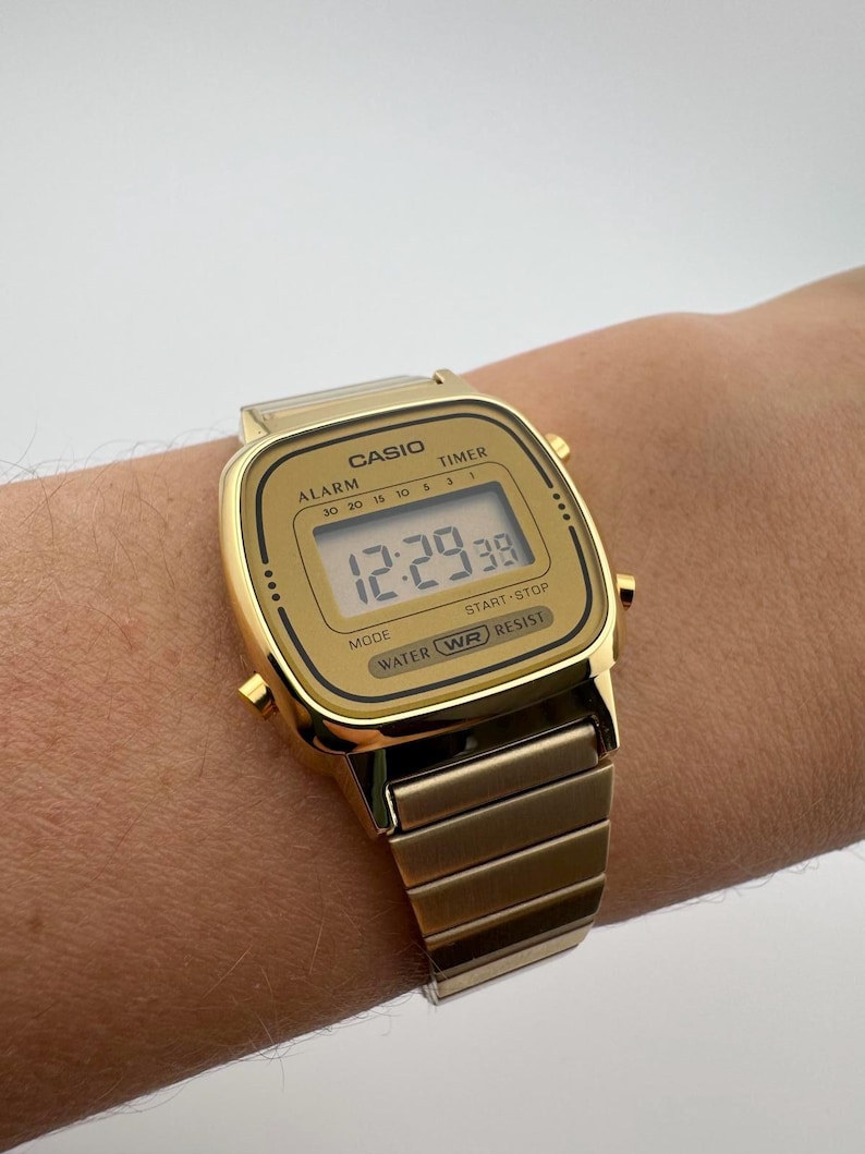 K&ouml;nnte beinhalten: Eine goldfarbene digitale Casio-Uhr mit rechteckigem Zifferblatt und passendem Metallarmband. Die Uhr zeigt die Zeit 12:29:38 an. Auf dem Zifferblatt stehen die W&ouml;rter "CASIO", "ALARM", "TIMER", "MODE", "START STOP" und "WATER WR RESIST".