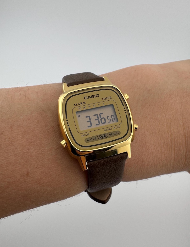 K&ouml;nnte beinhalten: Goldfarbene Casio Digitaluhr mit braunem Lederarmband. Die Uhr zeigt die Zeit 3:36:58 an. Das rechteckige Zifferblatt tr&auml;gt das Casio-Logo und Funktionsbezeichnungen. Das Armband ist braun.