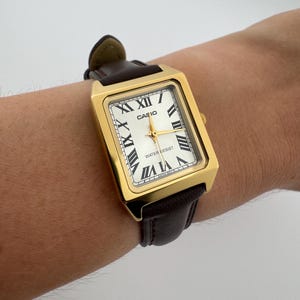 May include: Gold-tone rectangular Casio watch with a white face, black Roman numerals, and gold hands. The watch has a brown leather band. The watch face reads "CASIO" and "WATER RESIST".