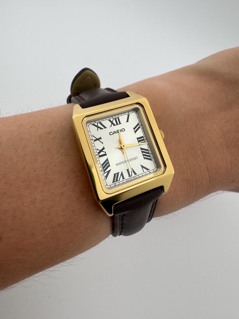 May include: Gold-tone rectangular Casio watch with a white face, black Roman numerals, and gold hands. The watch has a brown leather band. The watch face reads "CASIO" and "WATER RESIST".