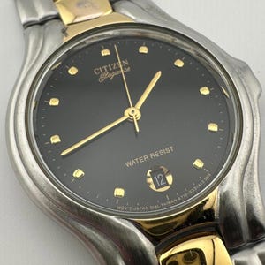 May include: A Citizen Elegance wristwatch featuring a black circular dial with gold-tone hour markers and hands. The watch has a two-tone silver and gold metal link bracelet. A date window displays the number 12 at the bottom of the dial.