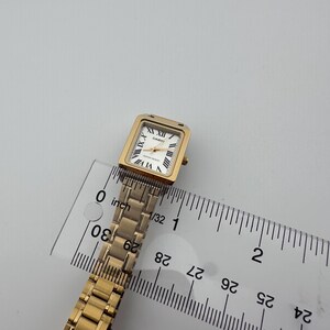 May include: A gold-toned Casio watch with a rectangular face and Roman numeral hour markers. The watch has a gold link band and is measured with a ruler. The watch face is approximately 0.75 inches wide.