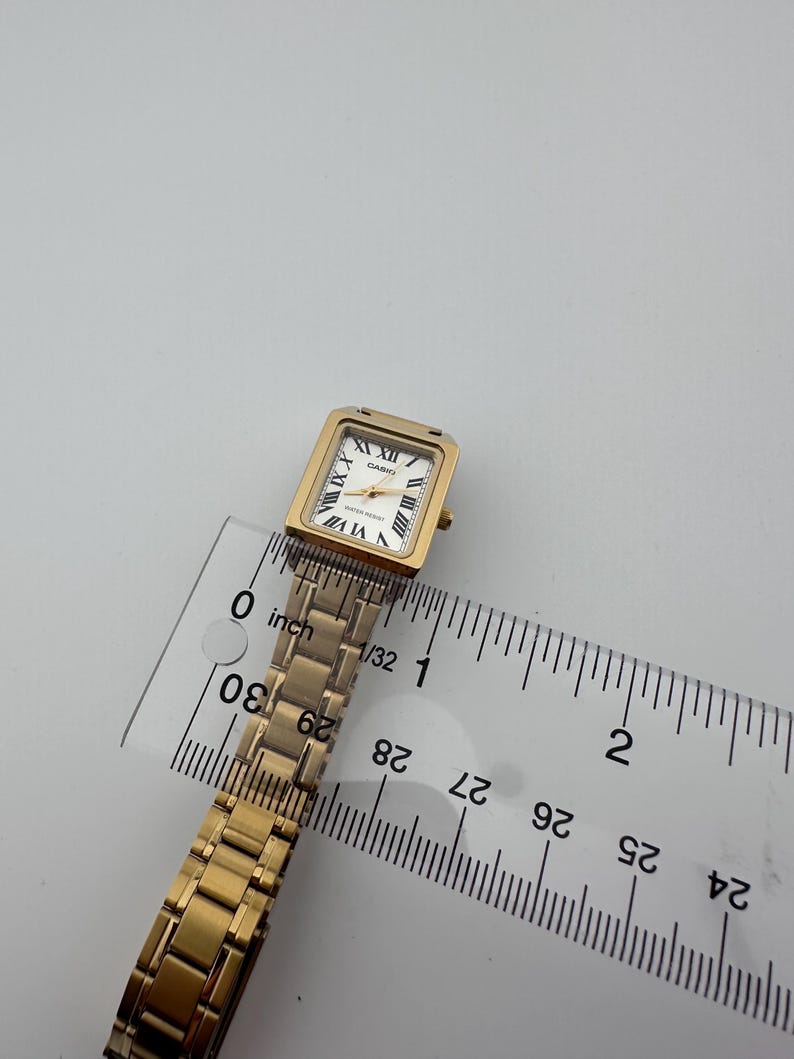 May include: A gold-toned Casio watch with a rectangular face and Roman numeral hour markers. The watch has a gold link band and is measured with a ruler. The watch face is approximately 0.75 inches wide.