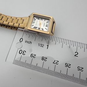 May include: A gold-toned Casio watch with a rectangular face and Roman numeral hour markers. The watch has a gold band and a white watch face. The watch is approximately 1 inch in length.