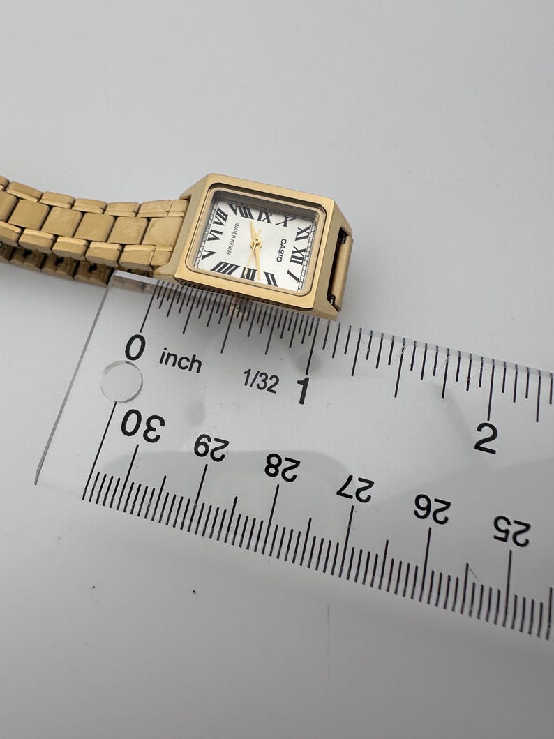 May include: A gold-toned Casio watch with a rectangular face and Roman numeral hour markers. The watch has a gold band and a white watch face. The watch is approximately 1 inch in length.
