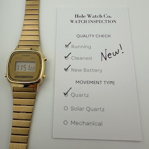 May include: A gold-toned Casio digital watch with a rectangular face and a matching metal band. The watch displays the time 11:51:22. A white card next to the watch reads "Hole Watch Co. WATCH INSPECTION" and lists quality checks.
