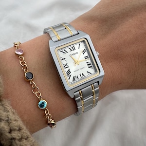 May include: Silver and gold-tone Casio watch with a rectangular face and Roman numeral hour markers. The watch is paired with a gold-tone bracelet featuring colorful gemstones. The watch band is silver with gold accents.