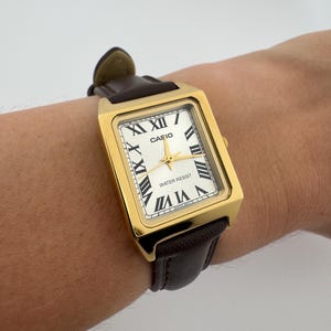 May include: A gold-toned rectangular Casio watch with a white face and black Roman numerals. The watch has gold hands and a brown leather band. The watch face reads "CASIO" and "WATER RESIST".