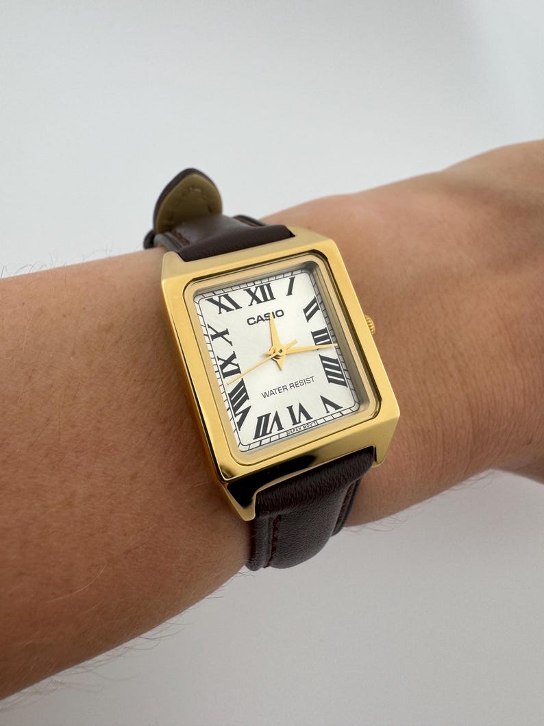 May include: A gold-toned rectangular Casio watch with a white face and black Roman numerals. The watch has gold hands and a brown leather band. The watch face reads "CASIO" and "WATER RESIST".