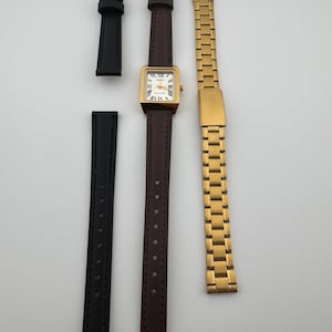 May include: A gold-toned square watch with a brown leather band, Roman numeral hour markers, and two additional watch bands: a black leather band and a gold metal link band. The watch face is white.