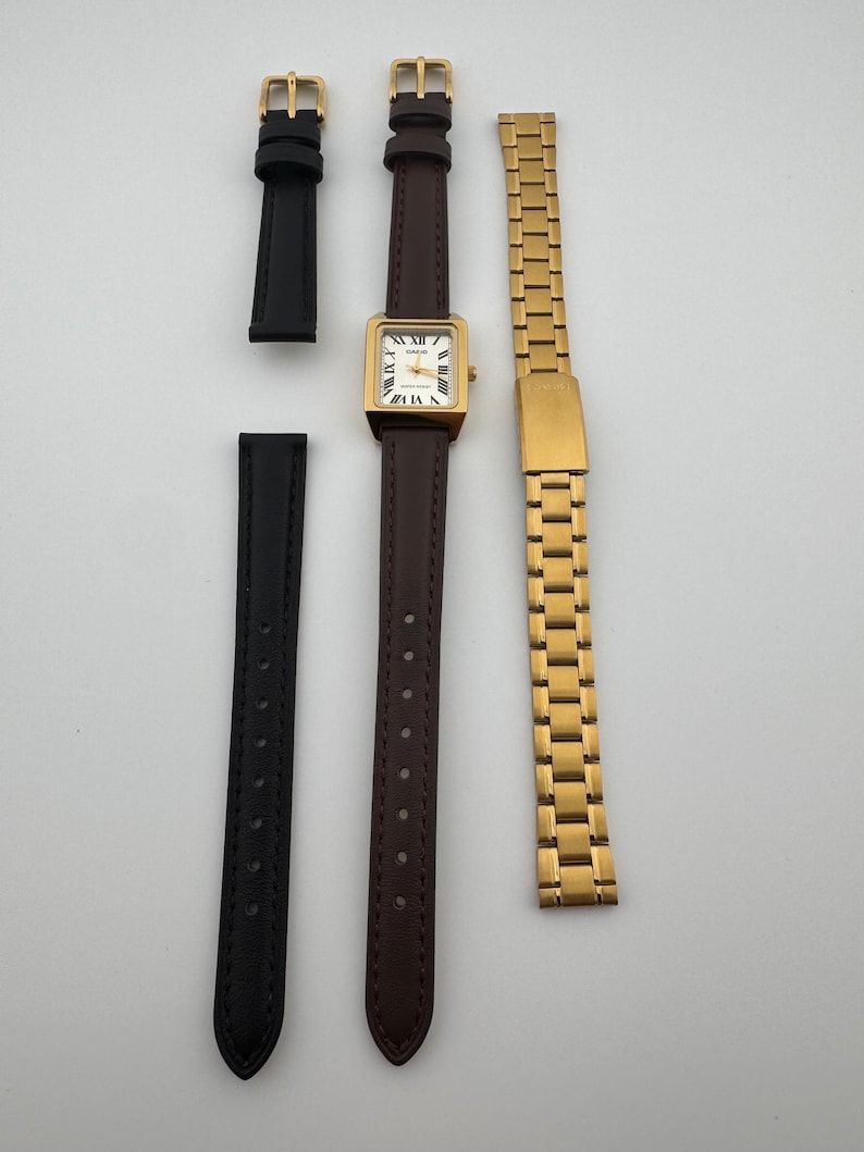 May include: A gold-toned square watch with a brown leather band, Roman numeral hour markers, and two additional watch bands: a black leather band and a gold metal link band. The watch face is white.