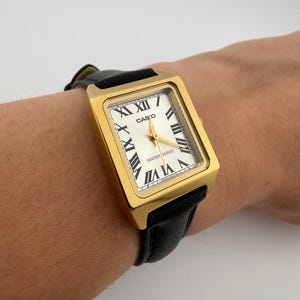 May include: A gold-toned Casio watch with a rectangular face and black leather band. The watch face is white with black Roman numerals and gold hands. The watch is water resistant.