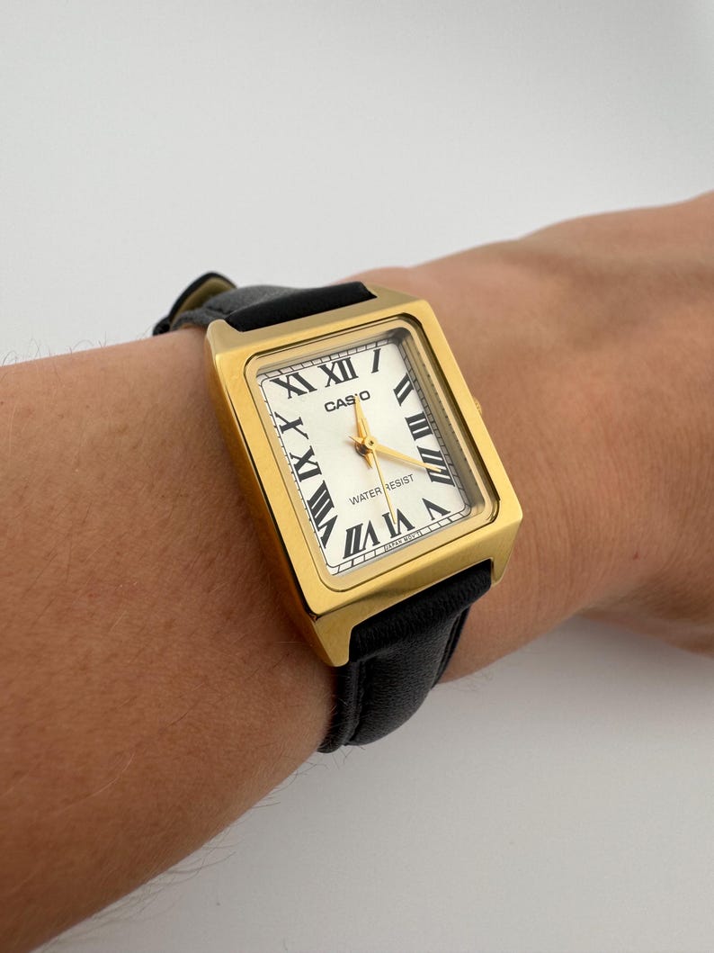 May include: A gold-toned Casio watch with a rectangular face and black leather band. The watch face is white with black Roman numerals and gold hands. The watch is water resistant.