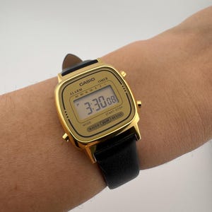 K&ouml;nnte beinhalten: Eine goldfarbene Casio Digitaluhr mit schwarzem Lederarmband. Das Zifferblatt zeigt die Uhrzeit 3:30:08 an. Die Uhr hat das Wort "CASIO" oben und "WATER WR RESIST" unten. Die Uhr befindet sich an einem Handgelenk.