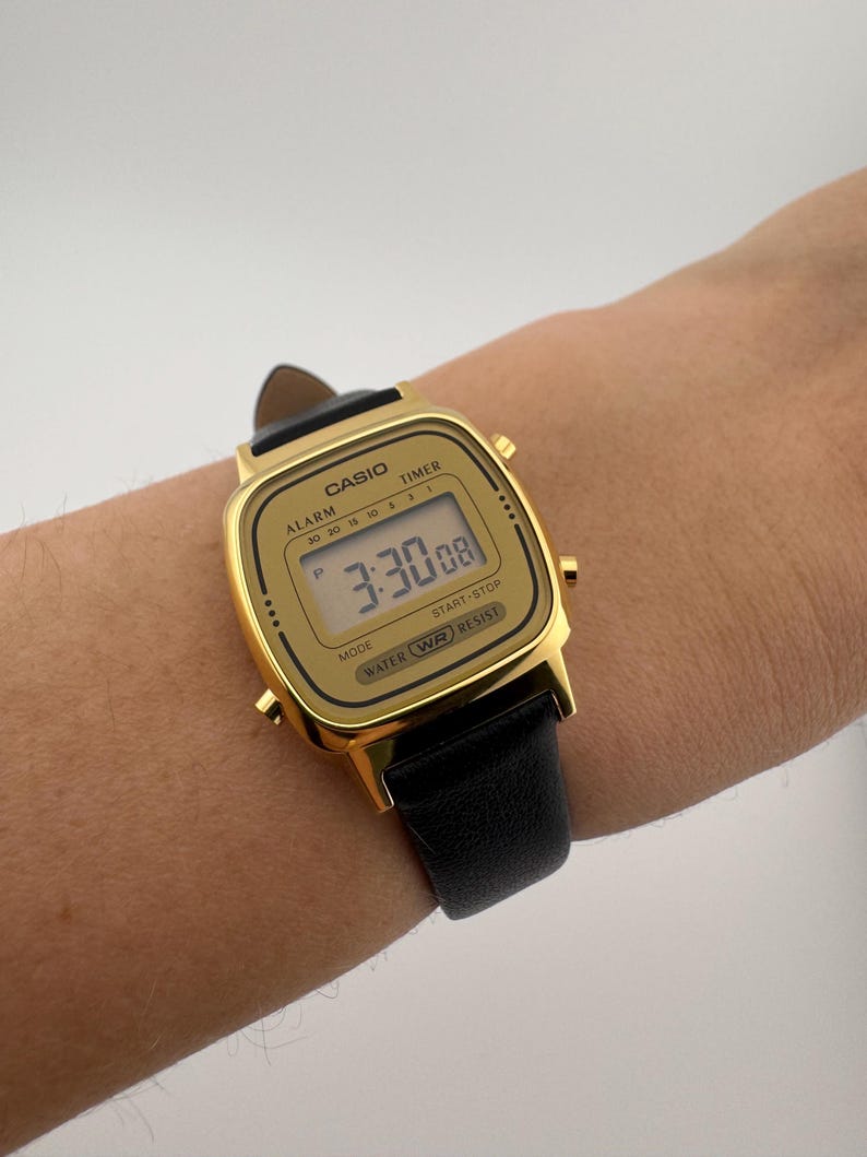 K&ouml;nnte beinhalten: Eine goldfarbene Casio Digitaluhr mit schwarzem Lederarmband. Das Zifferblatt zeigt die Uhrzeit 3:30:08 an. Die Uhr hat das Wort "CASIO" oben und "WATER WR RESIST" unten. Die Uhr befindet sich an einem Handgelenk.