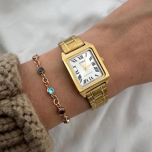 May include: A gold-toned rectangular wristwatch with a white face and Roman numeral hour markers is shown on a wrist. A gold bracelet with colorful gemstones is also visible. The watch band and case are gold-colored.