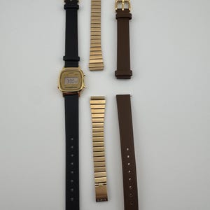 K&ouml;nnte beinhalten: Eine goldene Digitaluhr mit schwarzem Lederarmband und braunem Lederarmband. Ebenfalls enthalten sind zwei goldfarbene Metallarmb&auml;nder. Das Zifferblatt zeigt die Uhrzeit 3:00 Uhr an.