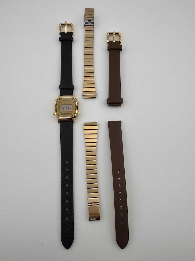K&ouml;nnte beinhalten: Eine goldene Digitaluhr mit schwarzem Lederarmband und braunem Lederarmband. Ebenfalls enthalten sind zwei goldfarbene Metallarmb&auml;nder. Das Zifferblatt zeigt die Uhrzeit 3:00 Uhr an.